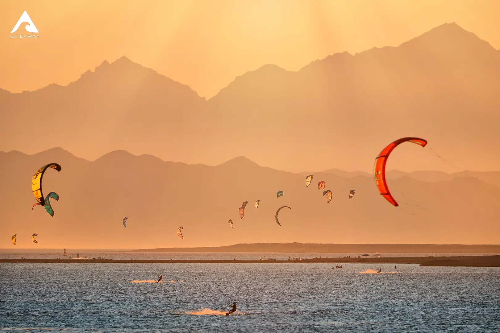 Kitesurferzy na wodzie o zachodzie słońca, barwne latawce i góry w tle - idealne wyjazdy kitesurfingowe z KiteAway.pl.