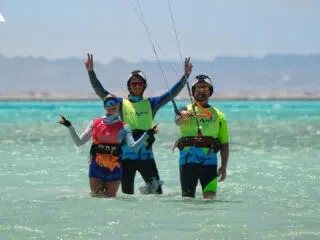 Trzy osoby na kursie kitesurfingu w kolorowych kamizelkach, z latawcami, stoją uśmiechnięte w wodzie podczas kite campu KiteAway.