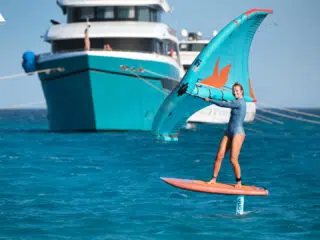 Uśmiechnięty kitesurfer na hydrofoilu i wingfoilu podczas kite tripu KiteAway; w tle jacht, idealne wakacje z kitesurfingiem.