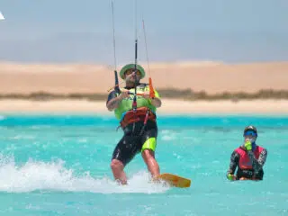 Kiteboarder na turkusowej wodzie podczas wyjazdu kitesurfingowego z KiteAway; obok osoby w piance, plaża w tle.