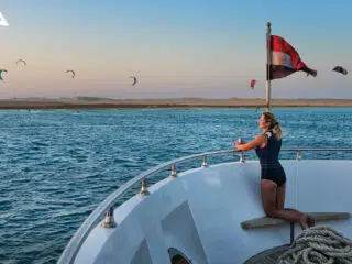 Kobieta w stroju kąpielowym na łodzi pod flagą KiteAway patrzy na kitesurferów; idealne wakacje z kitesurfingiem.