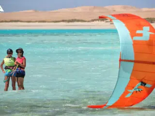 Dwie osoby w wodzie podczas kursu kitesurfingu KiteAway, szkolenie kite, kolorowy latawiec, piaszczyste wydmy w tle.