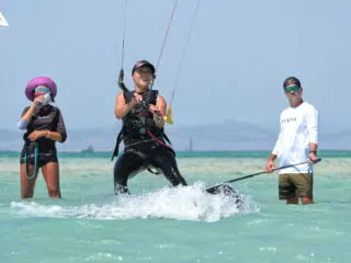 Uśmiechnięta kobieta na kursie kitesurfingu KiteAway, dwoje obserwujących, wakacje z kitesurfingiem, góry w tle.