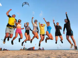 Grupa 8 osób skacze radośnie na plaży podczas wyjazdu kitesurfingowego KiteAway. Sprzęt i latawce w tle, słoneczna pogoda.