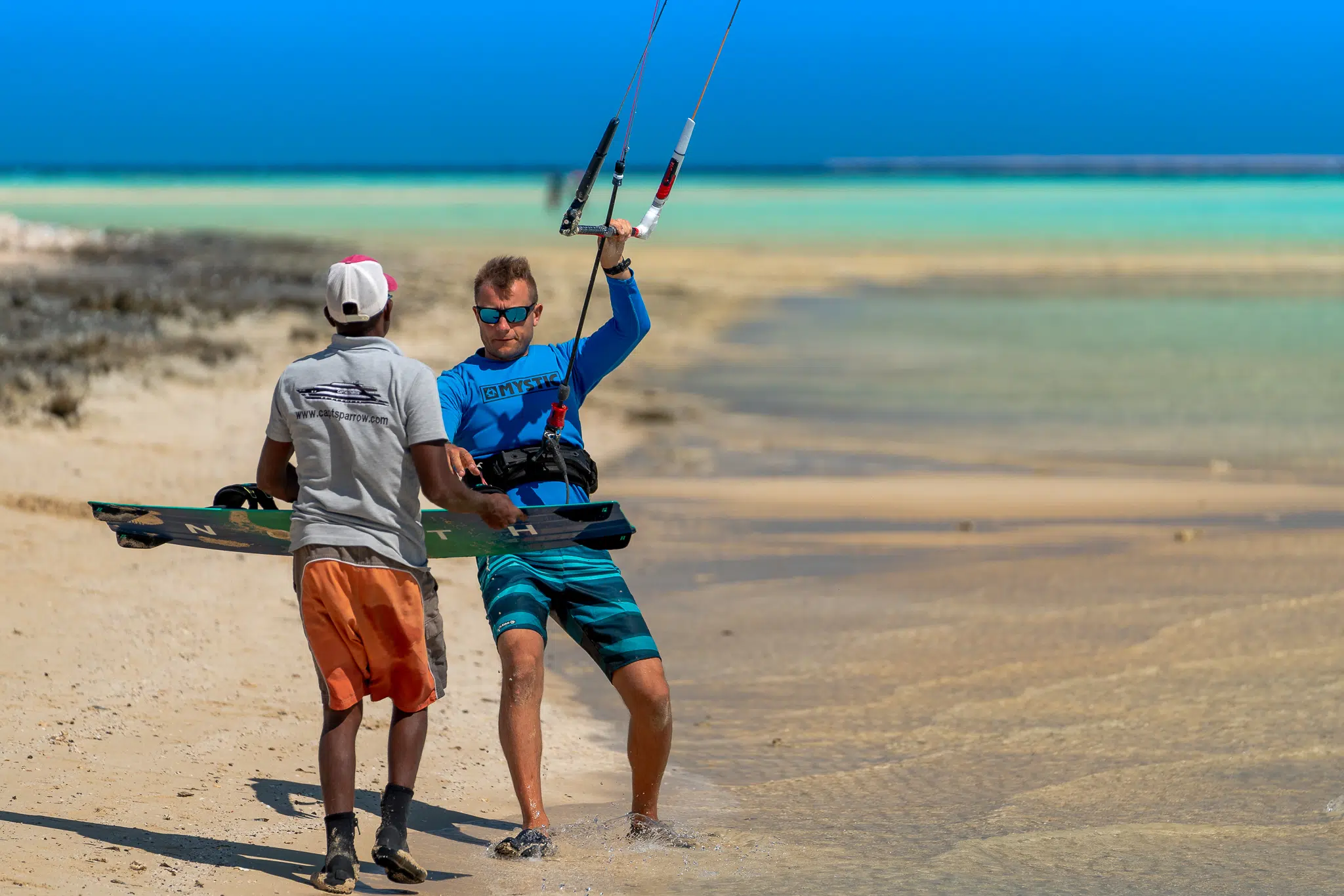 Dwóch mężczyzn na piaszczystej plaży z latawcem i deską, gotowych na wyjazd kitesurfingowy KiteAway pod błękitnym niebem.