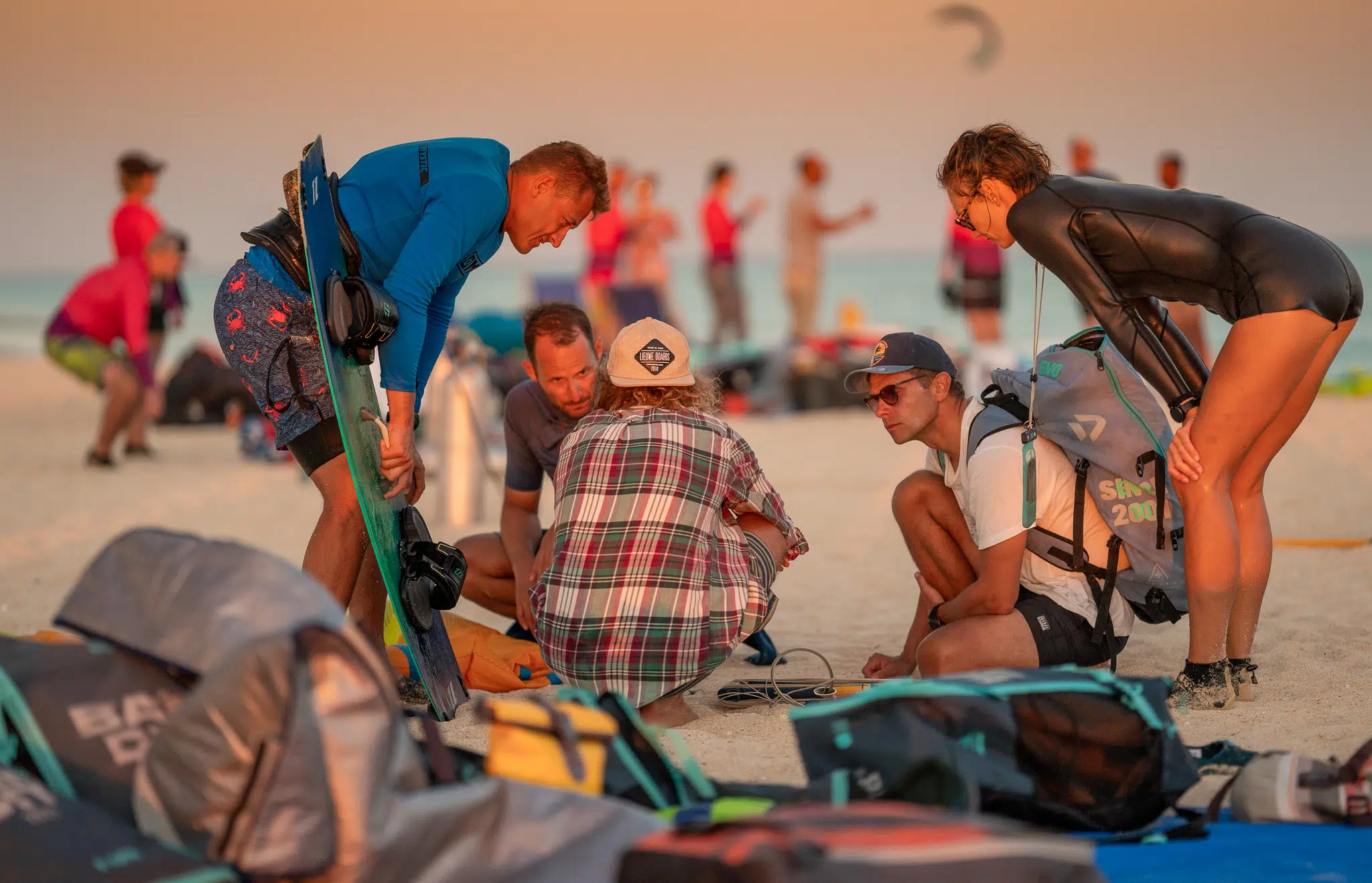 Uczestnicy wyjazdu kitesurfingowego KiteAway oglądają sprzęt na plaży; w tle kite camp, morze i zachód słońca.