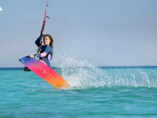 Kobieta w piance podczas kursu kitesurfingu unosi deskę nad krystaliczną wodą, dynamiczne wakacje z kitesurfingiem KiteAway.