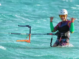 Uśmiechnięty kitesurfer na kite campie KiteAway, w kolorowym stroju i sprzęcie, stoi w turkusowej wodzie - gotowy na wyjazdy kitesurfingowe.