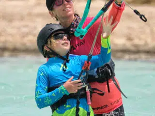 Instruktor KiteAway prowadzi kurs kitesurfingu z dzieckiem na płytkiej wodzie; w tle wydmy, idealne miejsce na wyjazdy kitesurfingowe.