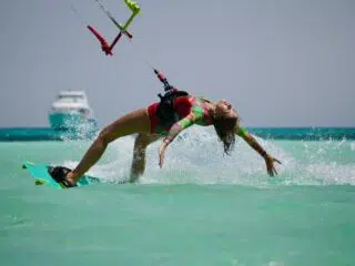 Kobieta w kolorowym stroju kitesurfinguje po turkusowej wodzie podczas wyjazdu KiteAway; w tle łódzki i wakacyjny kite trip.