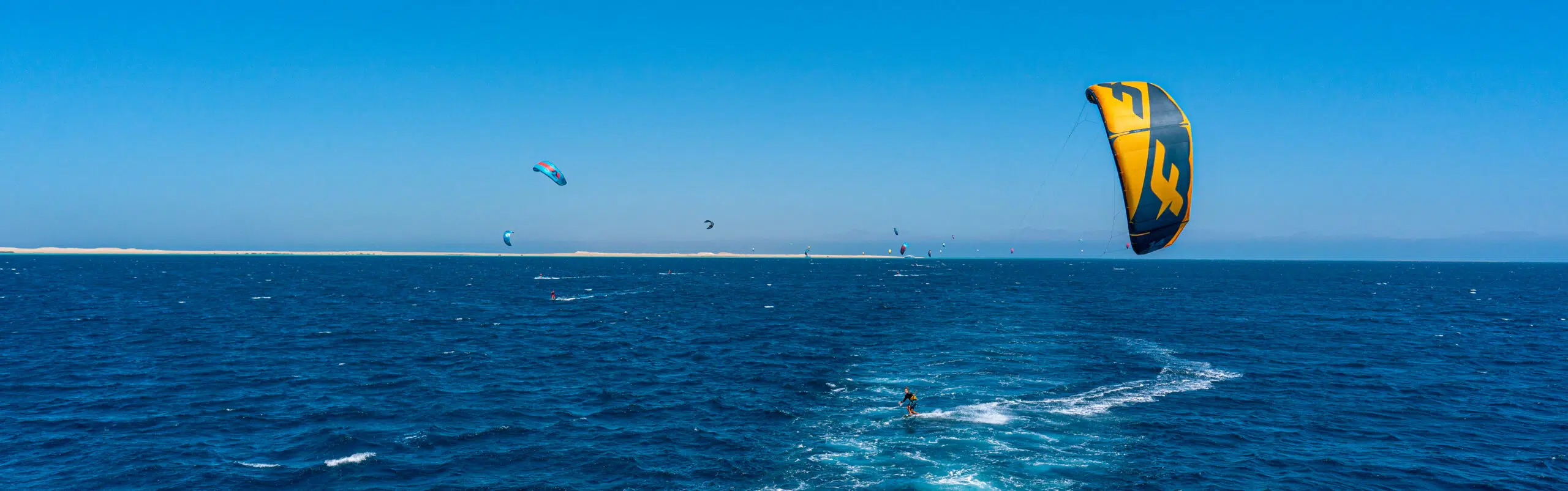 Kite camp KiteAway: szeroki akwen, kilku kitesurferów i duży żółto-niebieski latawiec, idealne warunki na wyjazdy kitesurfingowe.