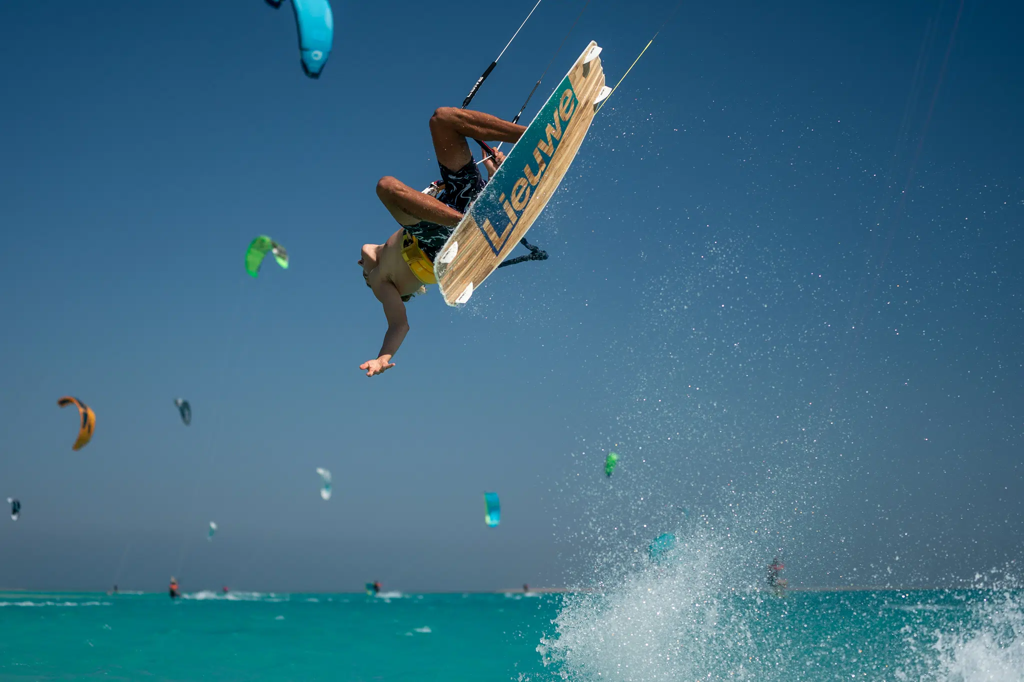 Kitesurfer wykonuje ewolucję nad turkusową wodą podczas wyjazdu kitesurfingowego KiteAway; w tle kolorowe latawce, wakacje z kitesurfingiem.