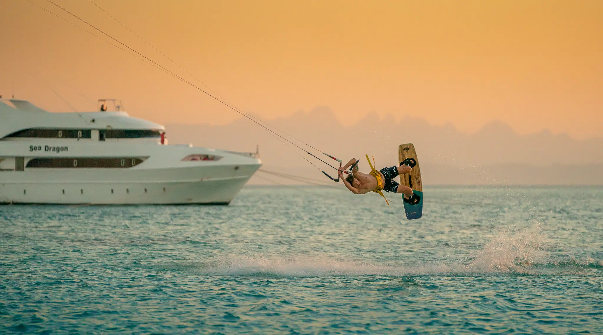 Kitesurfer wykonuje efektowny trick nad wodą blisko jachtu przy zachodzie słońca - idealne miejsce na wyjazdy kitesurfingowe z KiteAway.