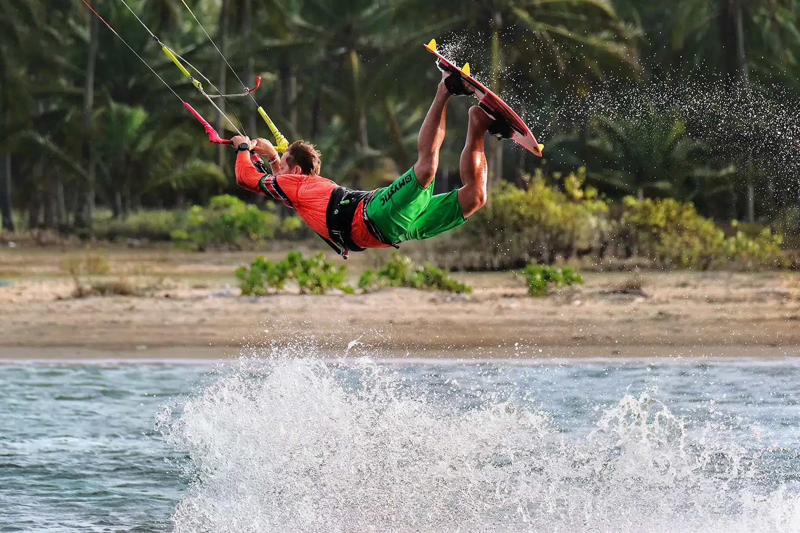 Kitesurfer w czerwonej koszulce nad wodą, egzotyczny kite spot z palmami, dynamiczna akcja - wakacje z kitesurfingiem KiteAway.
