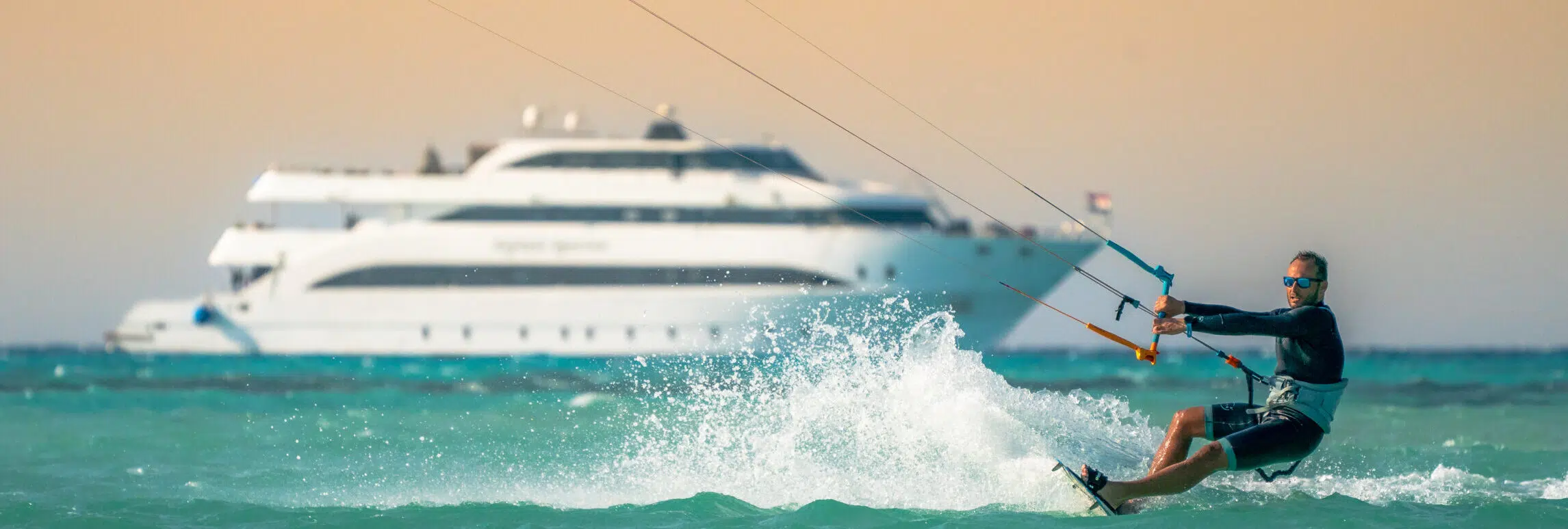 Mężczyzna w okularach pływa na kitesurfingu w turkusowej wodzie - wyjazdy kitesurfingowe z KiteAway, jacht w tle, zachód słońca.