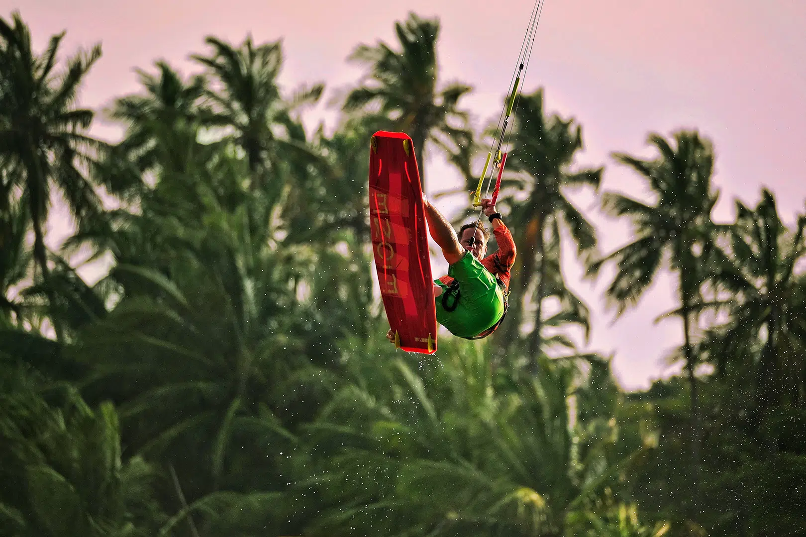 Kitesurfer w zielonej koszulce skacze z czerwonym kiteboardem na tle palm i różowego nieba podczas wyjazdu kitesurfingowego z KiteAway.