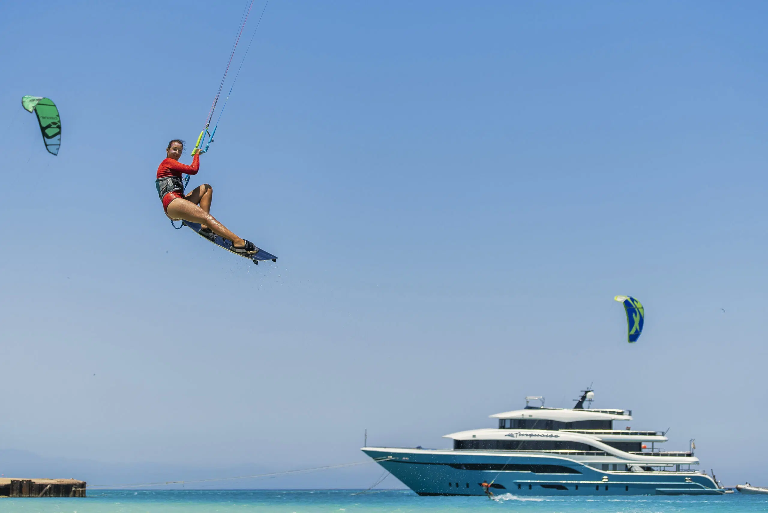 Kitesurfer w powietrzu nad turkusową wodą, w tle jacht; idealny dzień na wyjazdy kitesurfingowe z KiteAway.