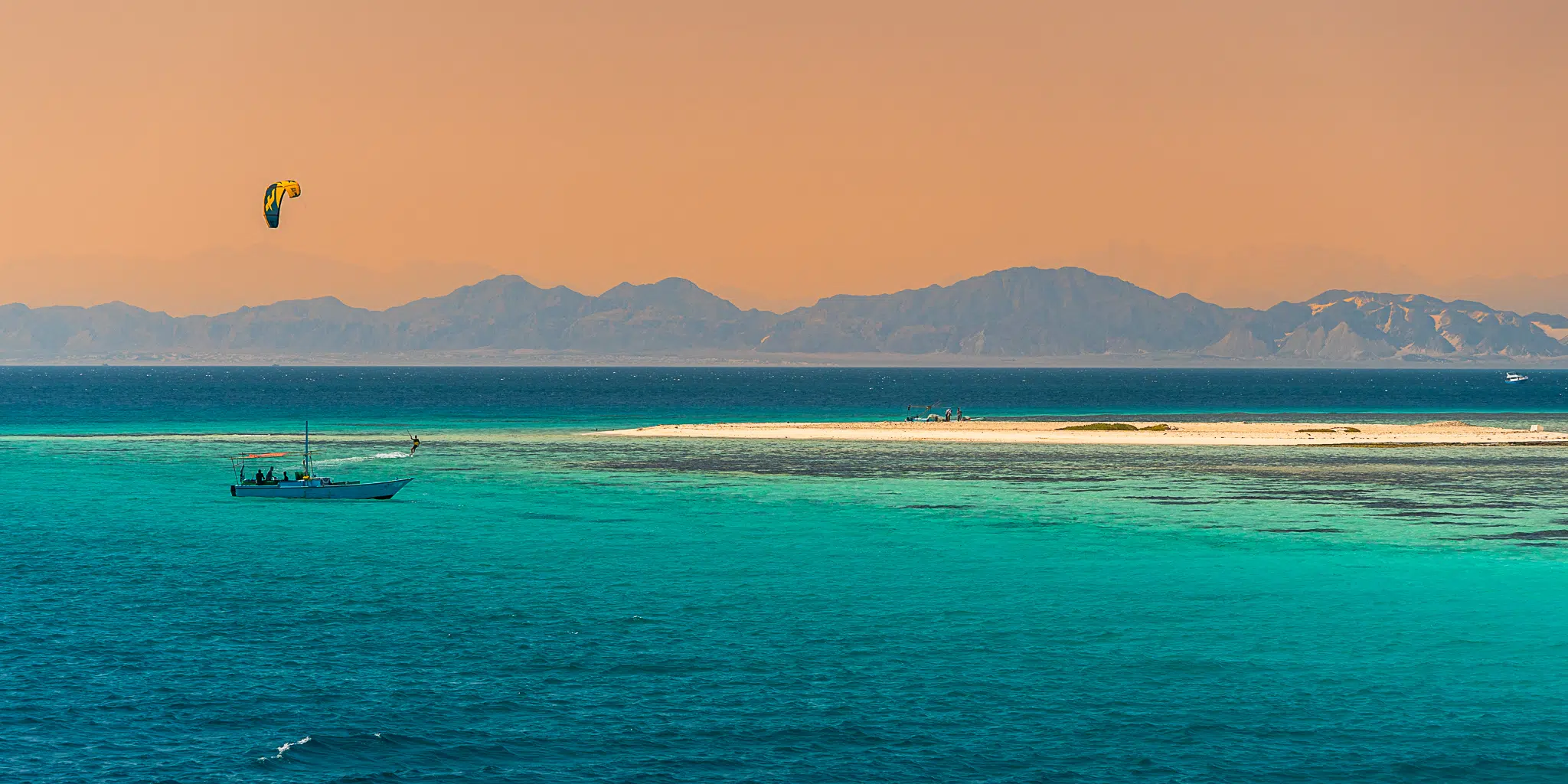 Mała łódź przy turkusowej wyspie i kitesurfer na tle gór-idealne miejsce na wyjazdy kitesurfingowe z KiteAway.
