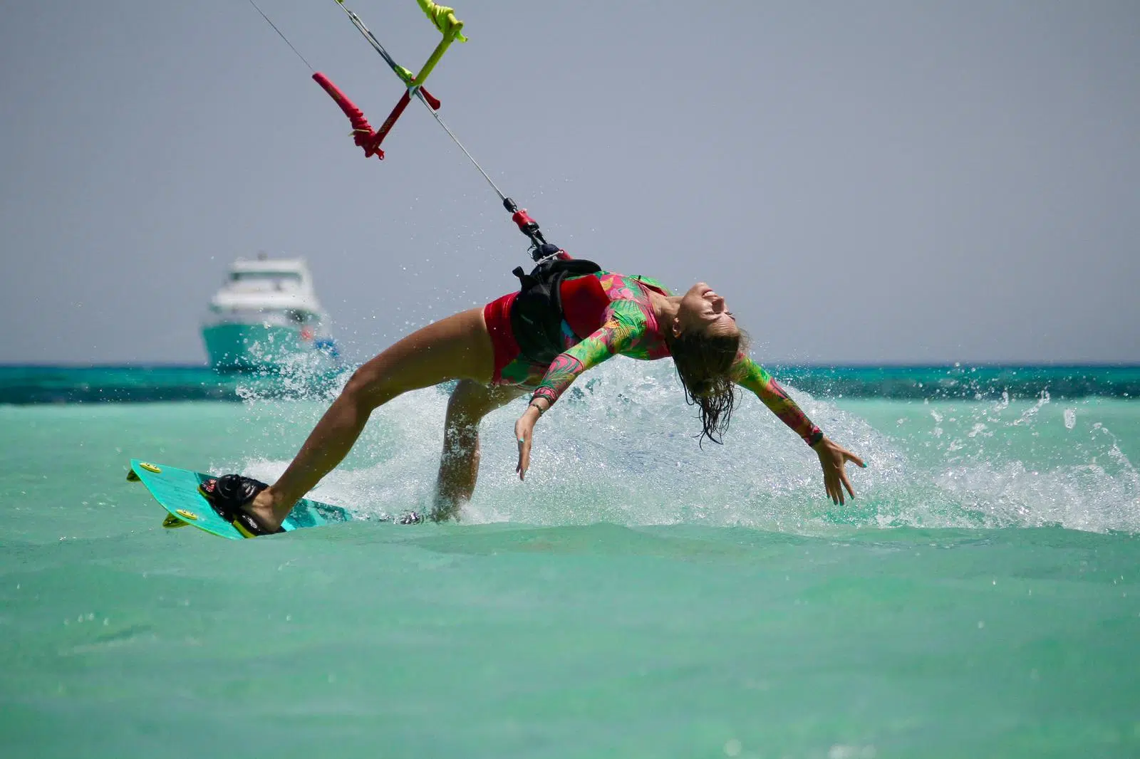 Kobieta w kolorowym piance wykonuje trick na turkusowej wodzie podczas wyjazdu kitesurfingowego z KiteAway; w tle łódź.