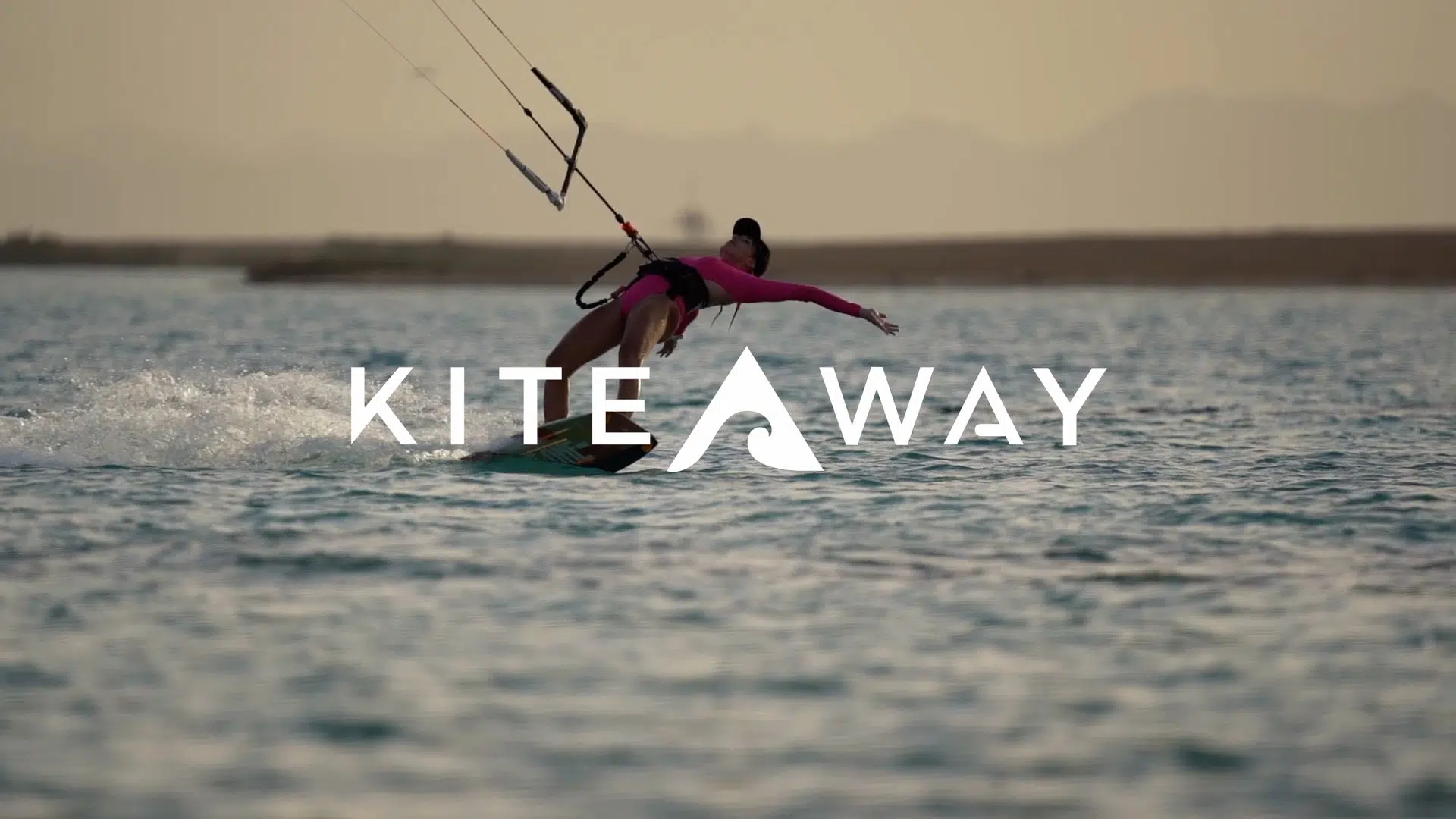 Osoba w różowym stroju kitesurfinguje o zachodzie słońca. Na zdjęciu logo KITEAWAY - wyjazdy kitesurfingowe i kite camp.