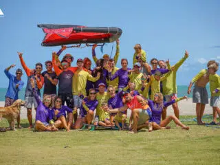 Uśmiechnięta grupa na trawie przy plaży podczas kite campu KiteAway, z czerwoną deską i psem. Idealne wyjazdy kitesurfingowe!.