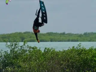Kitesurfer w piance wykonuje trick w powietrzu podczas wyjazdu kite z KiteAway.pl, w tle zieleń, woda i niebo.