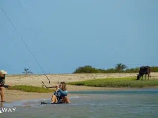 Kurs kitesurfingu KiteAway: instruktor na brzegu, kursant na desce w wodzie, obok pasąca się krowa, logo Kite Away, wakacje z kitesurfingiem.