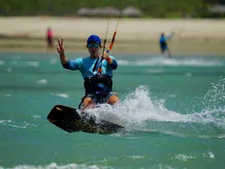 Uczestnik kite campu KiteAway kitesurfuje na turkusowej wodzie, robiąc znak pokoju; w tle plaża i wakacje z kitesurfingiem.