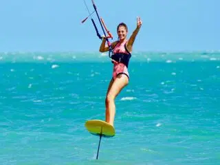 Uśmiechnięta kobieta kitefoiling na turkusowej wodzie, wakacje z kitesurfingiem z KiteAway, idealny kite trip pod błękitnym niebem.