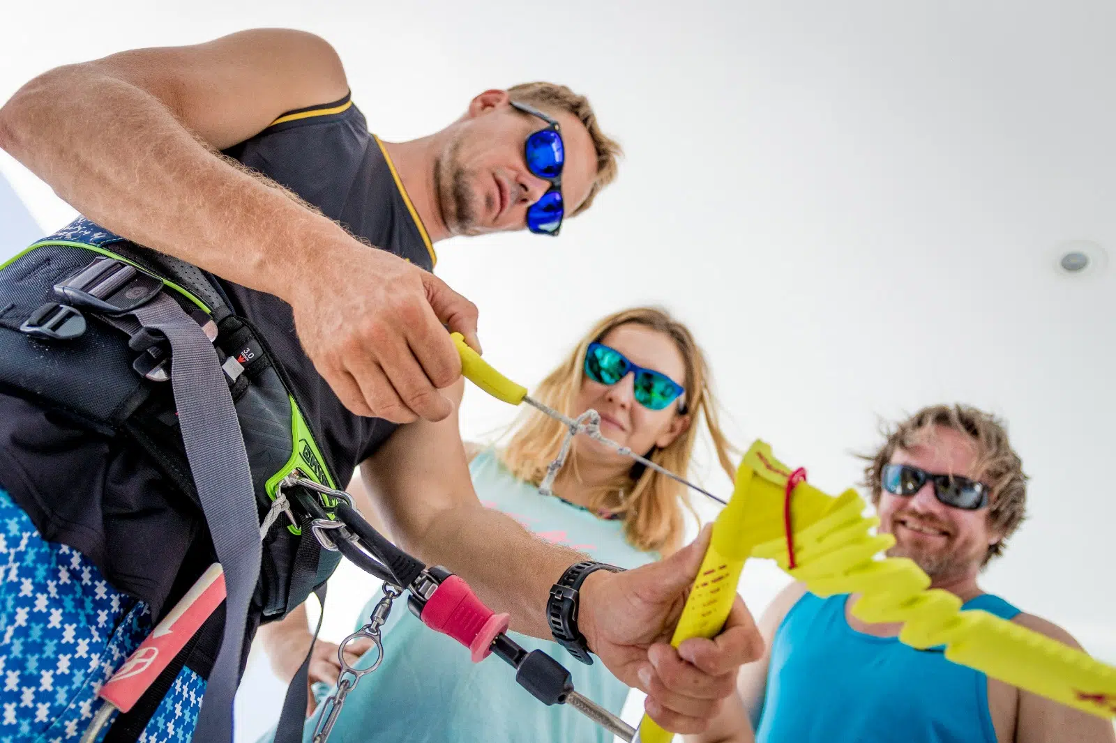 Trzy osoby w okularach i uprzężach przygotowują sprzęt na wyjazdy kitesurfingowe KiteAway, jasne tło, lato, kite trip.
