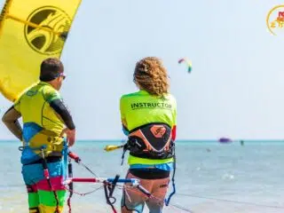 Instruktor i uczestnik kursu kitesurfingu KiteAway stoją w wodzie w El Gouna; na tle kitesurferzy, latawce, logo Red Sea Zone.