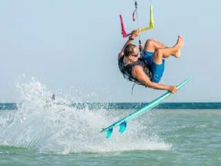 Kitesurfer w kamizelkach i niebieskich szortach robi trick nad wodą podczas wyjazdu kitesurfingowego KiteAway na słonecznej plaży.