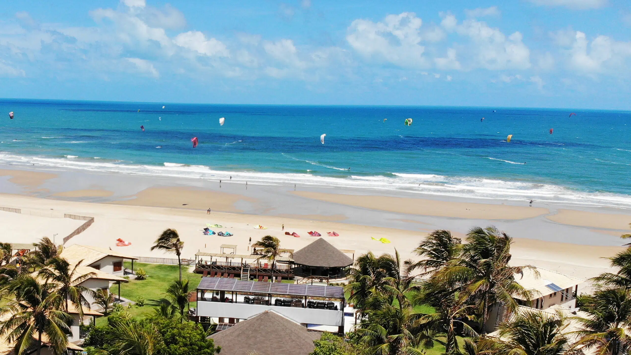 Piaszczysta plaża z palmami, budynkami i kitesurferami na niebieskim morzu. Kolorowe latawce KiteAway kite camp - wakacje z kitesurfingiem.