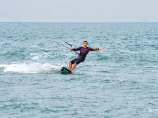 Uśmiechnięty uczestnik kursu kitesurfingu KiteAway płynie po spokojnej wodzie; w tle łódki i słoneczny kite spot.