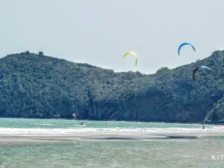 Kitesurferzy na turkusowej wodzie i kolorowe latawce, zielone wzgórze, logo KiteAway. Wyjazdy kitesurfingowe, kite camp.