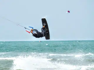 Kitesurfer wykonuje trik nad falami podczas wyjazdu KiteAway; w tle drugi uczestnik kite camp, słoneczna pogoda.