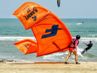 Osoba w różowym stroju szykuje pomarańczowego latawca na plaży podczas wyjazdu kitesurfingowego KiteAway; na tle kite campu.