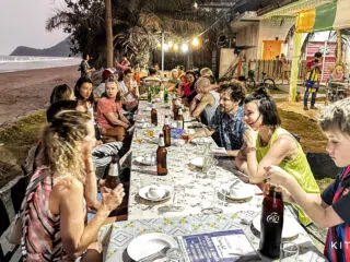 Grupa osób przy stole na plaży podczas zachodu słońca, kolorowe lampki; klimatyczny kite camp KiteAway, wakacje z kitesurfingiem.