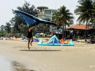 Osoba w czerni skacze z niebieskim latawcem na plaży KiteAway - wakacje z kitesurfingiem, kolorowe kite'y, palmy.