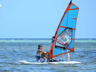Osoba w okularach i kolorowych szortach siedzi na desce na płytkiej wodzie podczas wakacji z kitesurfingiem KiteAway.
