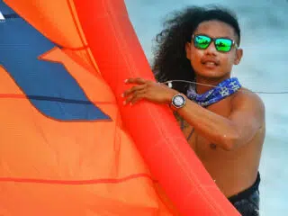 Uśmiechnięty kitesurfer z długimi lokami i bandaną trzyma pomarańczowy latawiec na plaży podczas wyjazdu KiteAway - kite camp.