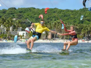 Para kitesurferów trzyma się za ręce na wodzie podczas wyjazdu KiteAway; w tle palmy, plaża i kolorowe latawce, wakacje z kitesurfingiem.
