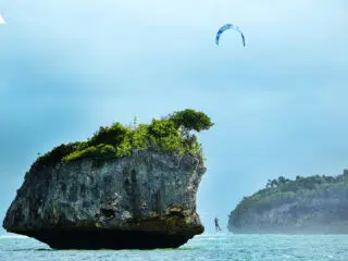 Kitesurfer płynący przy skalistej wyspie z zielenią, pod pochmurnym niebem - idealny spot na wyjazdy kitesurfingowe KiteAway.