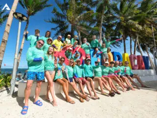 Uczestnicy kite campu KiteAway w turkusowych koszulkach na plaży w Cabarete podczas wyjazdu kitesurfingowego, palmy i nieba.