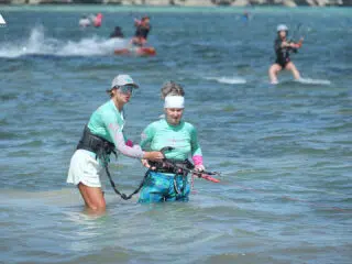 Instruktorka prowadzi kurs kitesurfingu z uczestniczką w płytkiej wodzie, na tle kite campu KiteAway i innych kitesurferzy.