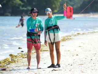 Instruktor KiteAway prowadzi kurs kitesurfingu na plaży; uczestnicy kite tripu trenują na tle wody i drzew.