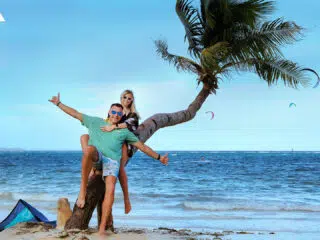 Uśmiechnięta para na palmie przy plaży, słońce, fale i kitesurferzy - idealny klimat na wyjazdy kitesurfingowe z KiteAway.