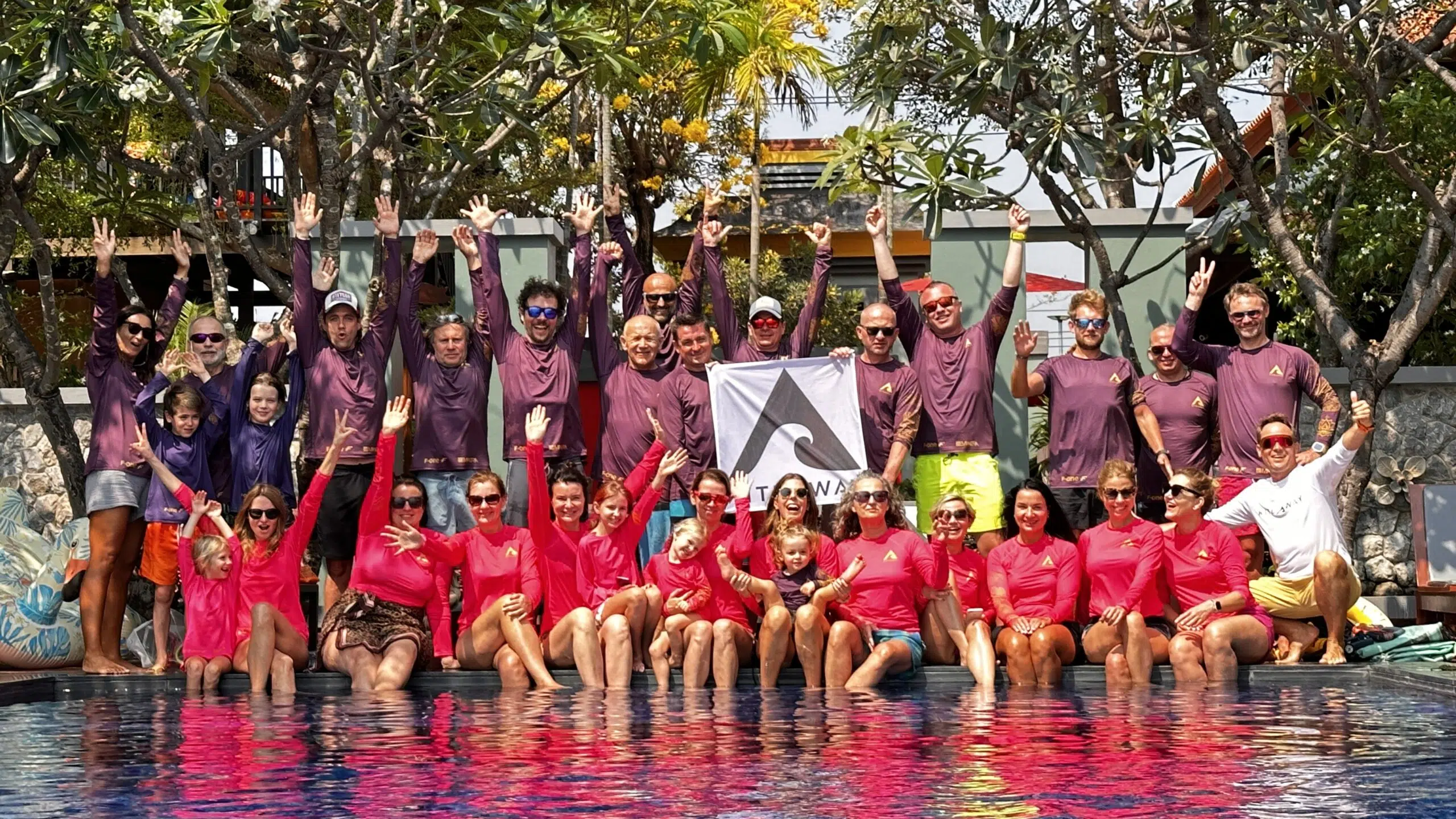 Uczestnicy wyjazdu kitesurfingowego KiteAway w kolorowych koszulkach świętują przy basenie pod banerem "kite camp".