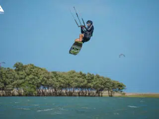 Kitesurfer w powietrzu nad wodą, z drzewami i błękitem nieba w tle - dynamiczna scena z wyjazdów kitesurfingowych KiteAway.