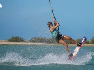 Kitesurfer w turkusowej kamizelce unosi się nad wodą na wyjeździe KiteAway; w tle piaszczysta plaża i błękitne niebo.